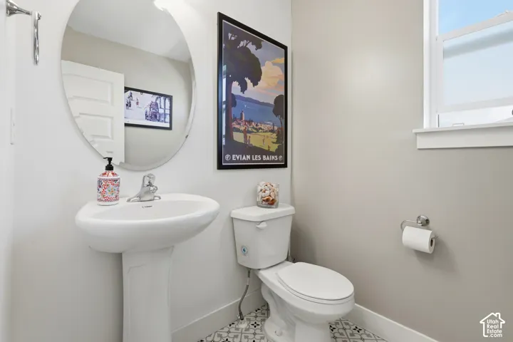 Pedestal sink, wall-mounted mirror, white toilet, single-pane window, and patterned tile flooring