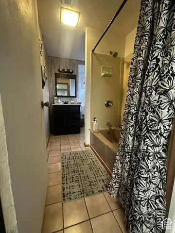 Ceramic tile flooring, vanity with dark wood-finish cabinetry, rectangular mirror, three-light vanity fixture, and a built-in bathtub with shower