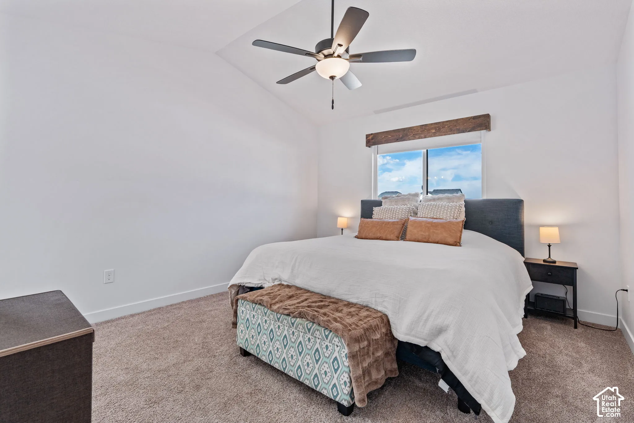 Vaulted ceiling, ceiling fan with integrated lighting, window with wood-finish header, carpeted flooring, and a built-in bed frame with an upholstered headboard