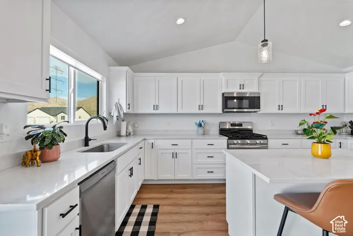 Vaulted ceiling with recessed lighting, white cabinetry with black hardware, stainless steel appliances, light-toned countertops, and wood-finish flooring
