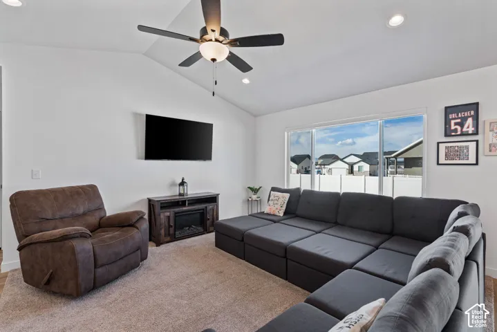 Spacious recreation room featuring a sectional layout with ample seating, ceiling fan with integrated lighting, wall-mounted flat-screen television, dark wood-finish media console, neutral carpet flooring, and expansive windows overlooking the neighborhoo