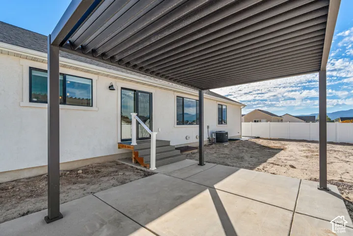 Expansive concrete patio with a modern louvered pergola, sliding glass door access, exterior lighting, and a white stucco facade