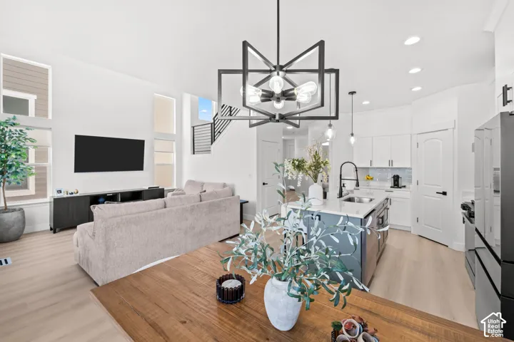 Open-concept living space with wood-finish flooring, a contemporary black geometric chandelier, a kitchen island with a white countertop and integrated sink, white kitchen cabinetry with a tile backsplash, and a modern staircase with metal balusters