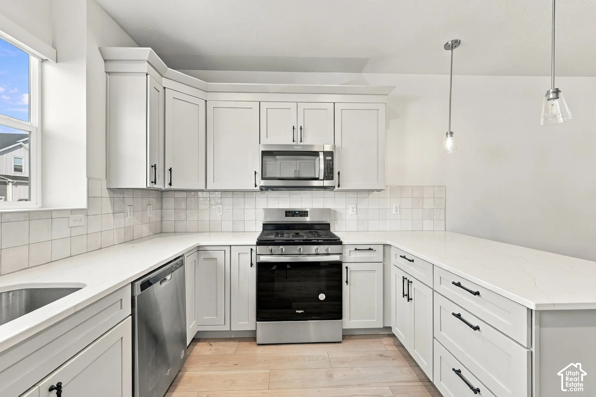 White shaker cabinetry with black hardware, stainless steel appliances, light-toned wood-finish flooring, white solid surface countertops, and a white square tile backsplash