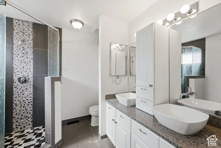 Dual vessel sinks with polished chrome faucets, granite-style countertop, white cabinetry with polished chrome pulls, and a glass-enclosed shower with mosaic tile accents