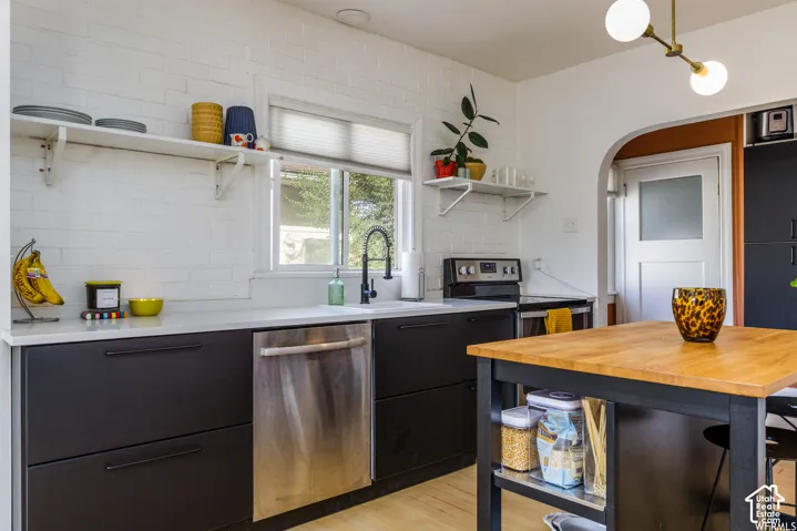 Kitchen featuring a white brick wall, dark flat-panel cabinetry, a stainless steel dishwasher, a light-toned wood-finish island, and a modern ceiling light fixture