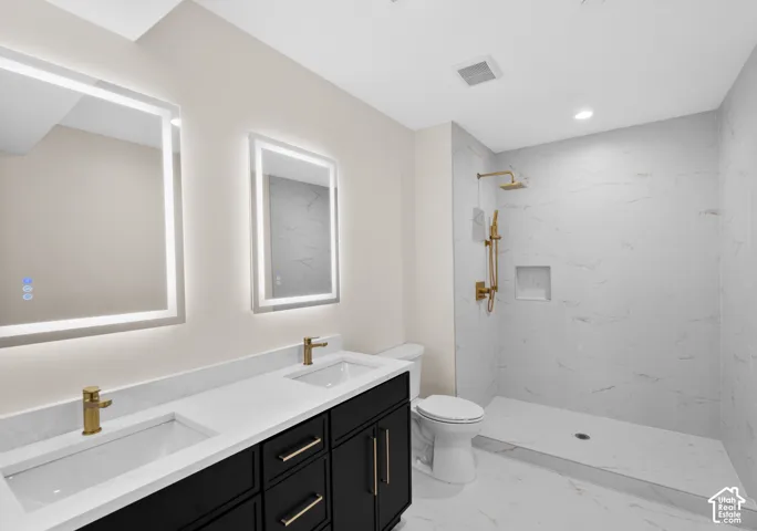 Dual illuminated mirrors, a double vanity with stone countertops, and a walk-in shower with gold-tone fixtures and a recessed niche