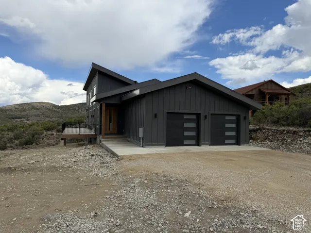 Contemporary exterior with dark vertical siding, a two-car garage featuring modern doors with horizontal window panels, an elevated wood-finish deck with black railings, and a prominent wood-finish front entry door