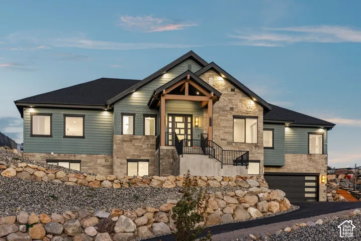Multi-level residence featuring stone and horizontal siding exterior, prominent covered entry with wood beams, black asphalt driveway, and integrated two-car garage with modern frosted glass panels