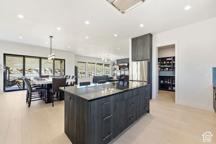 Open-concept living space featuring light wood-finish flooring, a large kitchen island with dark cabinetry, stainless steel refrigerator, and a walk-in pantry