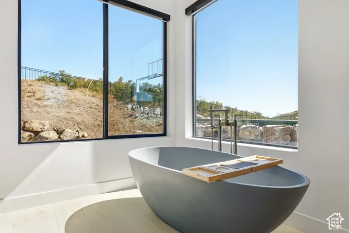 Freestanding soaking tub with a floor-mounted fixture, large windows, and light wood-finish flooring