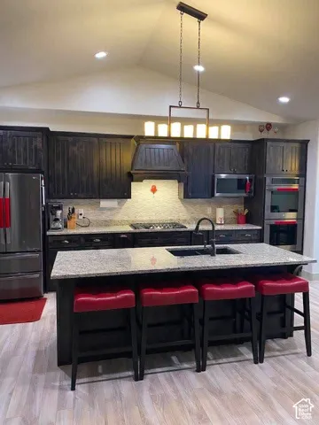Vaulted ceiling with recessed lighting, dark wood-finish cabinetry, stainless steel appliances, central island with sink, and light wood-finish flooring