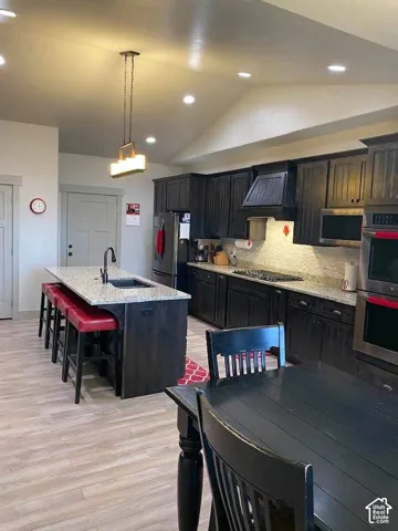 Kitchen featuring dark wood cabinetry, light-toned wood-finish flooring, a central island with an integrated sink, recessed lighting, and a vaulted ceiling
