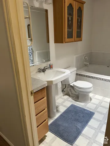 Pedestal sink with chrome fixtures, tiled bathtub surround, wall-mounted cabinetry with glass inserts, and vanity with wood-finish drawers