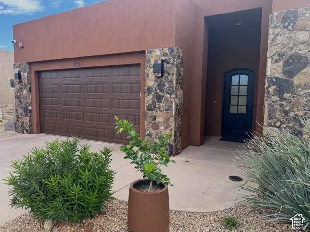Stucco exterior with stone accents, wood-look garage door, covered entry, arched entry door with glass panels, and concrete walkway