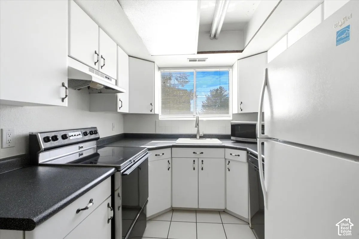 White cabinetry with dark hardware, dark countertops, white tile flooring, integrated sink with a gooseneck faucet, and a large window
