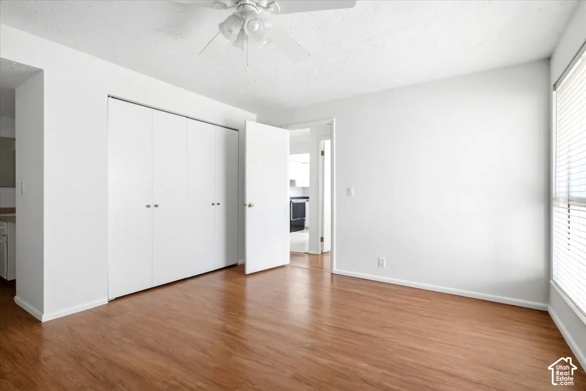 Wood-finish flooring, white walls, white ceiling fan, large windows with blinds, and built-in closet with white bifold doors