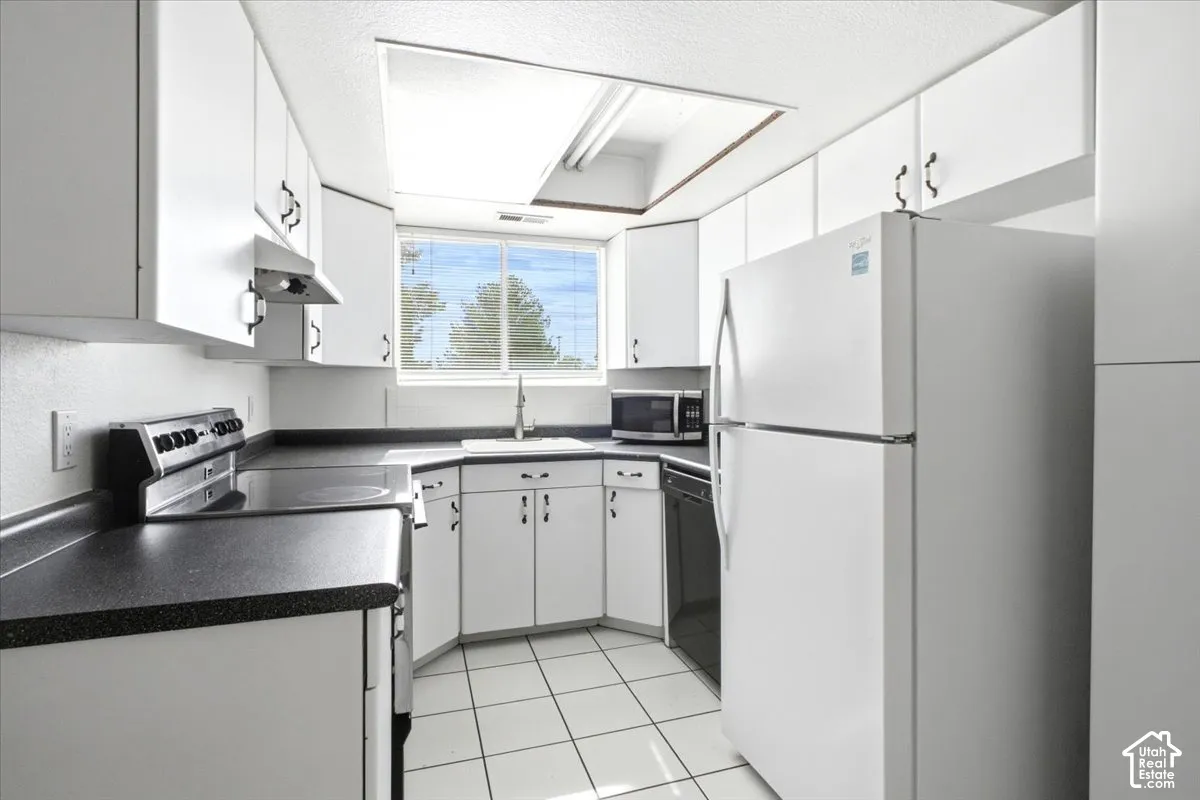 White cabinetry with dark hardware, black countertops, white floor tiling, and a window above the sink