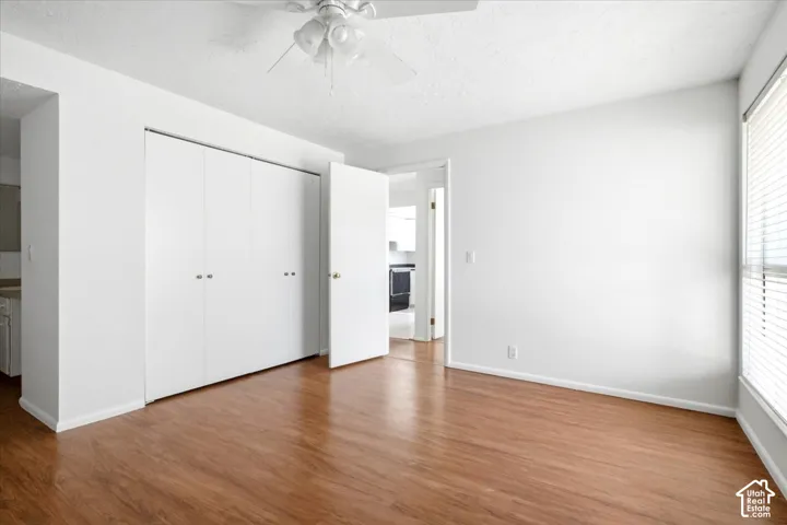 Wood-finish flooring, white walls, white ceiling fan, large windows with blinds, and built-in closet with white bifold doors