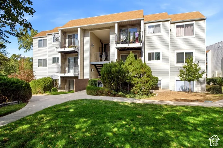 Three-story apartment building with light-colored siding, multiple balconies, exterior stairs, and surrounding landscaping