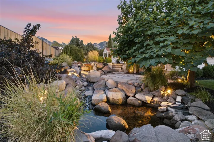 Expansive backyard featuring a multi-level rock waterfall, stone patio, mature trees, and landscape lighting