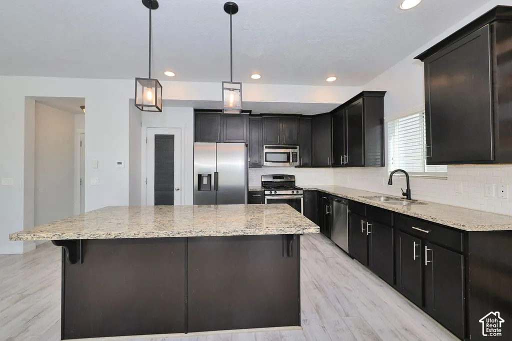 Kitchen featuring dark cabinetry, stone countertops, stainless steel appliances, a center island with pendant lighting, and wood-finish flooring