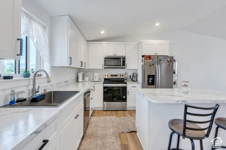 White cabinetry with dark hardware, stainless steel appliances, a kitchen island with seating, marbled countertops, and wood-finish flooring