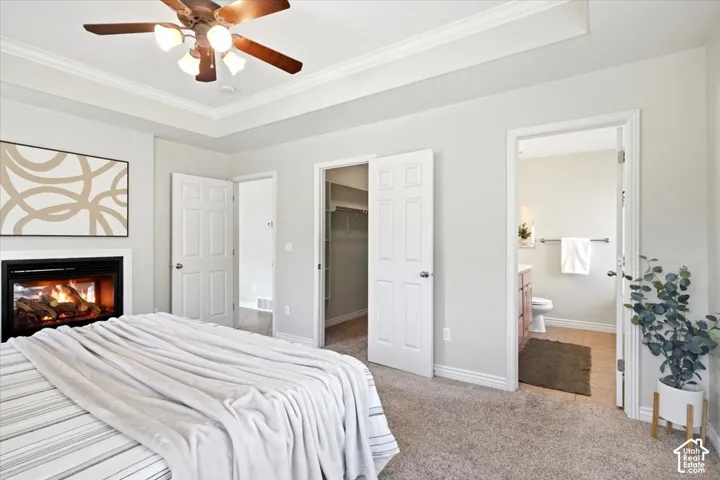 Recessed ceiling with crown molding, ceiling fan, built-in fireplace with black surround, light-toned carpet flooring, and an en-suite bathroom with a wood-finish vanity and tiled floor