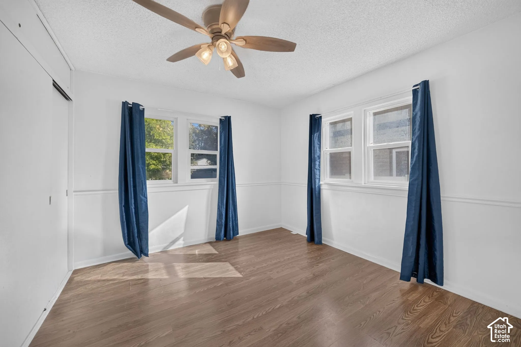 Wood-finish flooring, two large windows, white paneled walls, and a ceiling fan with integrated lighting