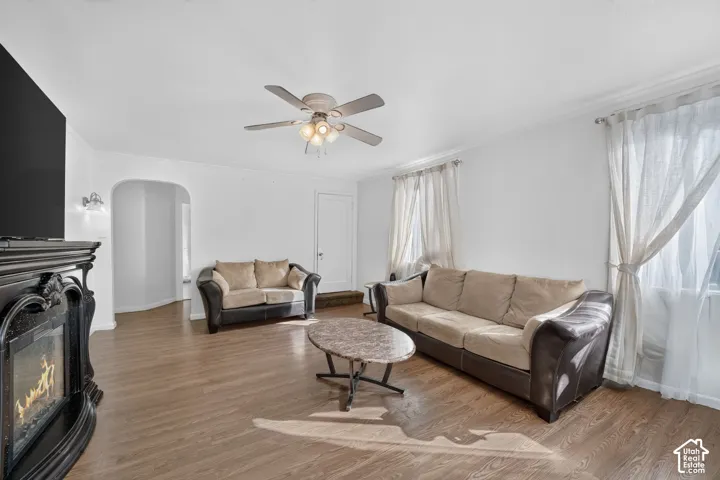 Wood-finish flooring, ceiling fan with integrated lighting, wall-mounted light fixtures, arched doorway, and a decorative fireplace mantel