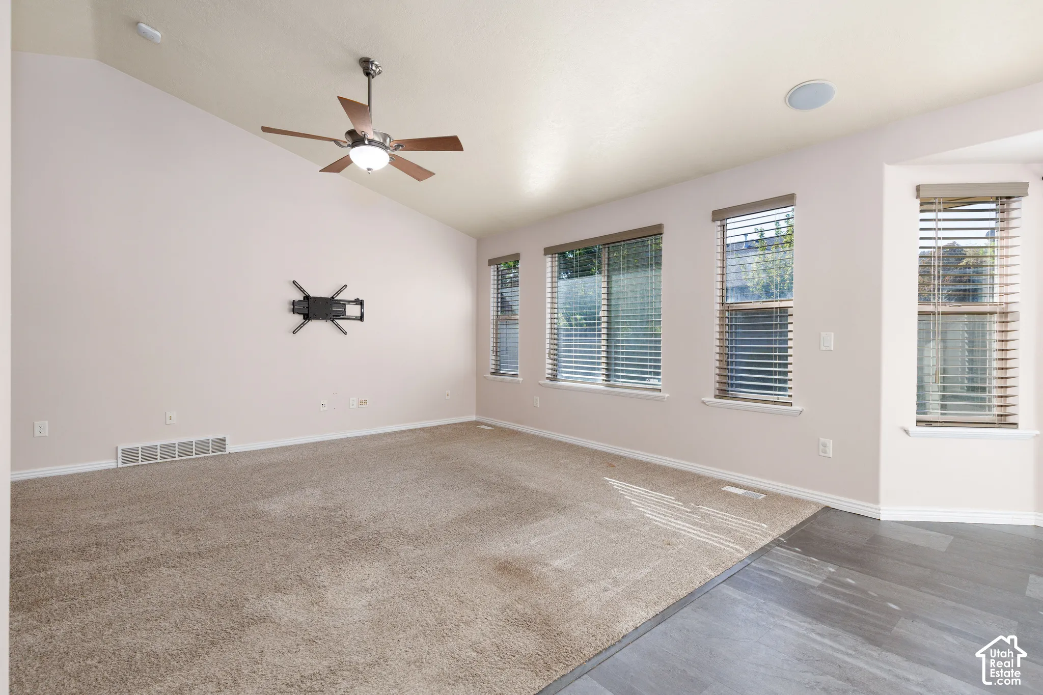 Vaulted ceiling, ceiling fan with integrated lighting, multiple windows with blinds, built-in ceiling speaker, and mixed flooring with carpet and wood-finish