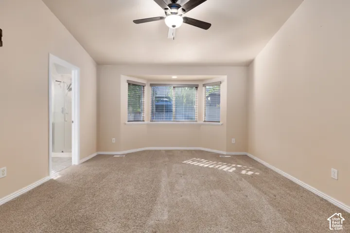Carpeted room with a bay window, ceiling fan with light fixture, and an entry to a bathroom with a glass-enclosed shower