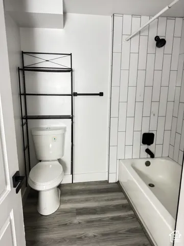 White subway tile shower surround with matte black fixtures, white bathtub, wood-finish flooring, and white walls