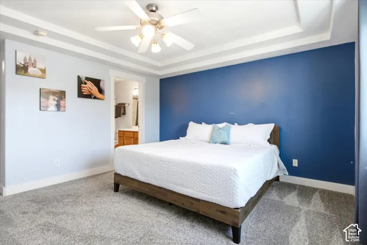 Tray ceiling with integrated lighting, ceiling fan with light kit, textured carpet flooring, painted accent wall, and an en-suite bathroom with a wood-finish vanity