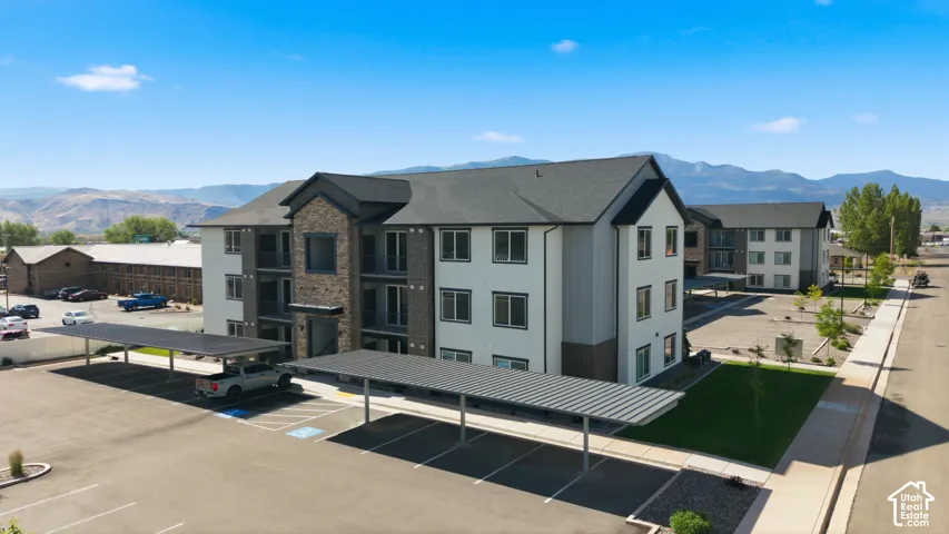 Contemporary multi-story building featuring stone and siding exterior, covered parking structures, paved parking lot, and manicured landscaping