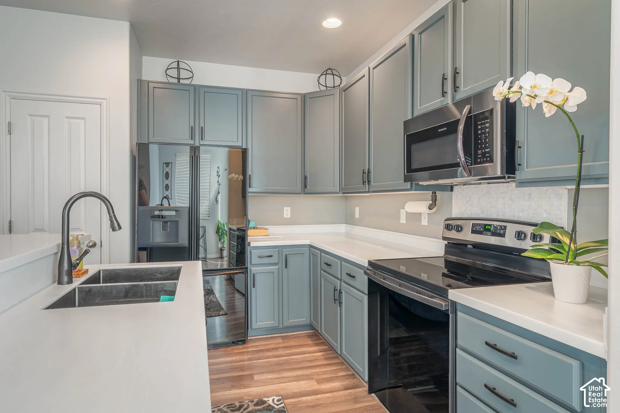 Light blue cabinetry with matte black hardware, stainless steel appliances, solid surface countertops, a double basin sink with a gooseneck faucet, and wood-finish flooring