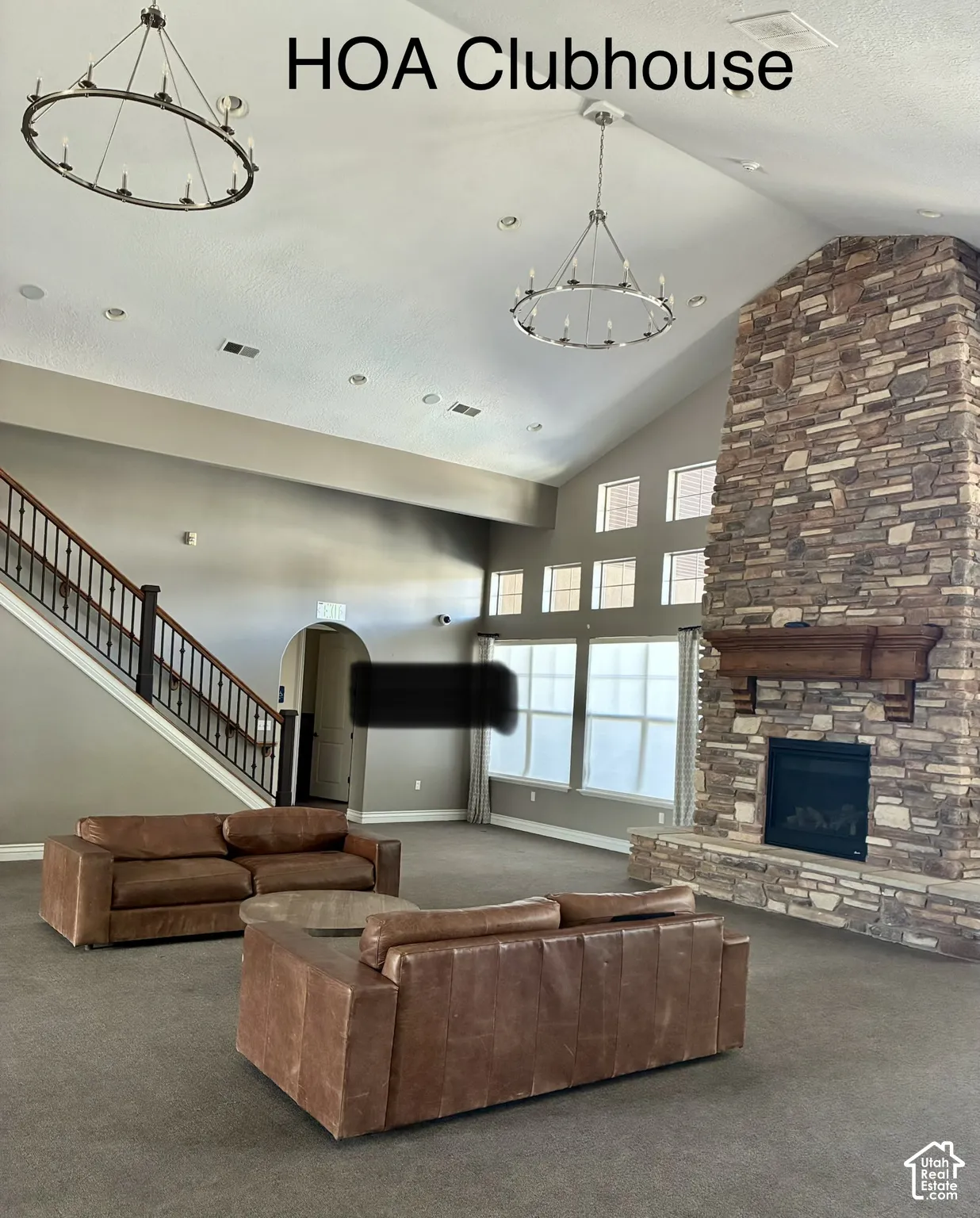 Vaulted ceiling, stone-clad fireplace with wood mantel, multiple windows, staircase with wood handrail, and chandelier lighting
