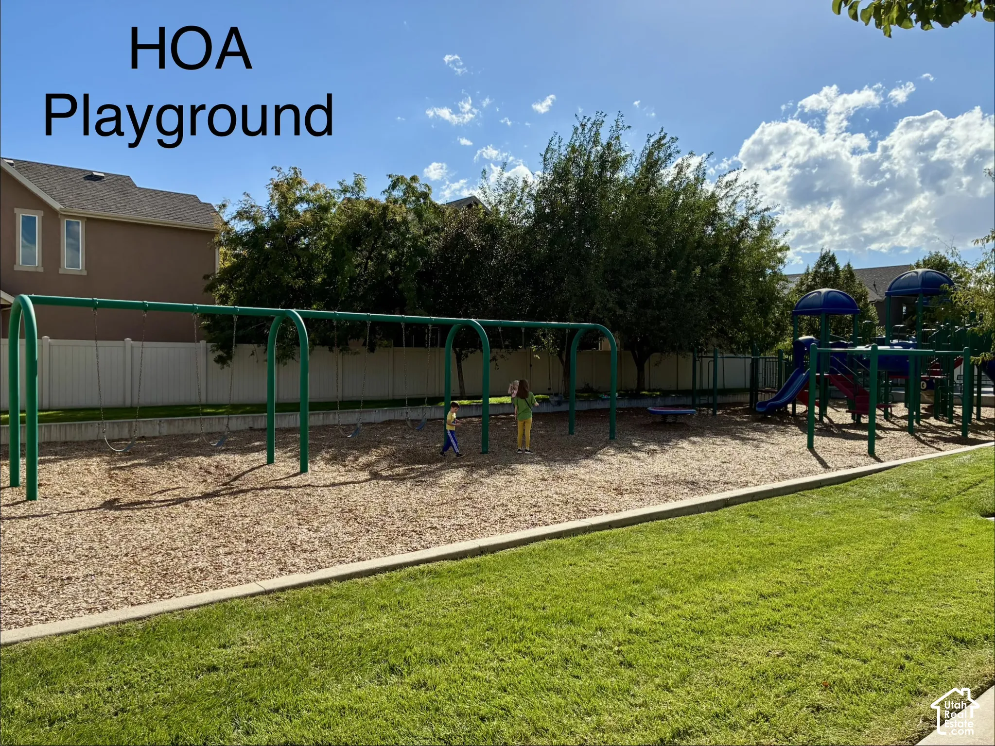 Community playground featuring a swing set, play structure with slides, and wood chip surfacing, bordered by manicured lawn
