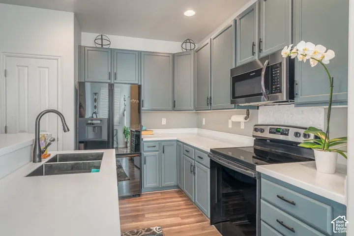 Light blue cabinetry with matte black hardware, stainless steel appliances, solid surface countertops, a double basin sink with a gooseneck faucet, and wood-finish flooring