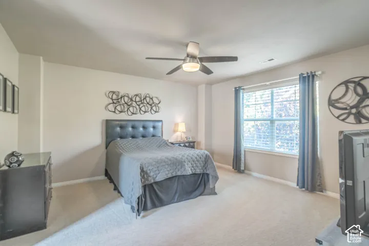 Light-colored carpeting, ceiling fan with integrated lighting, window with blinds, and upholstered headboard