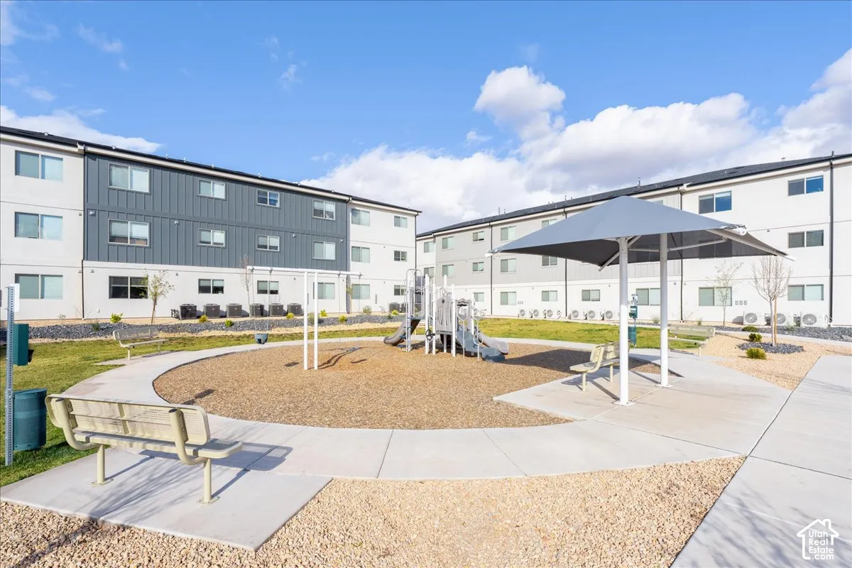 Community green space featuring a shaded play structure, playground area, concrete pathways, and a seating bench