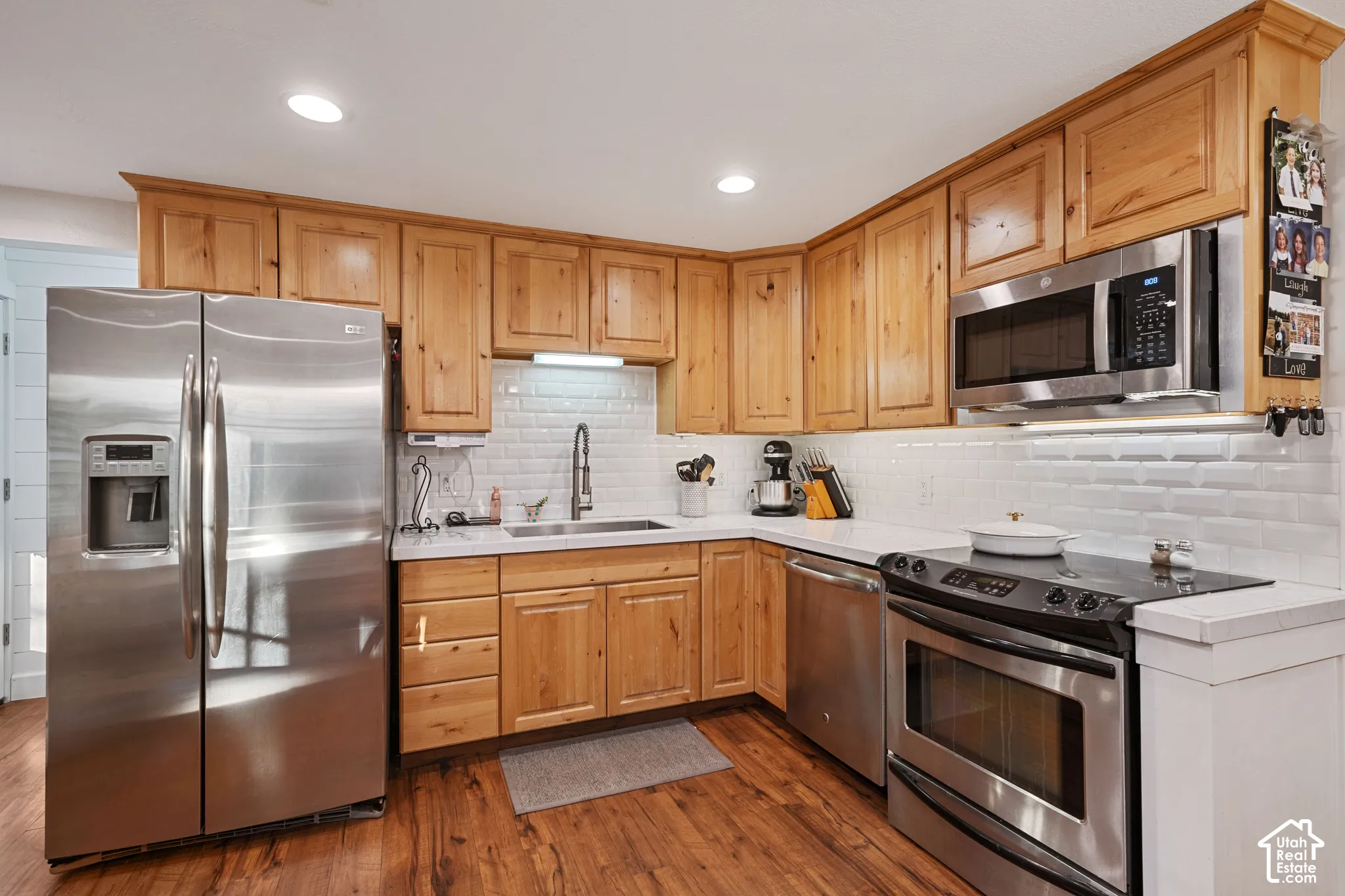 Kitchen featuring wood-finish flooring, light wood cabinetry, stainless steel appliances, white subway tile backsplash, and white countertops