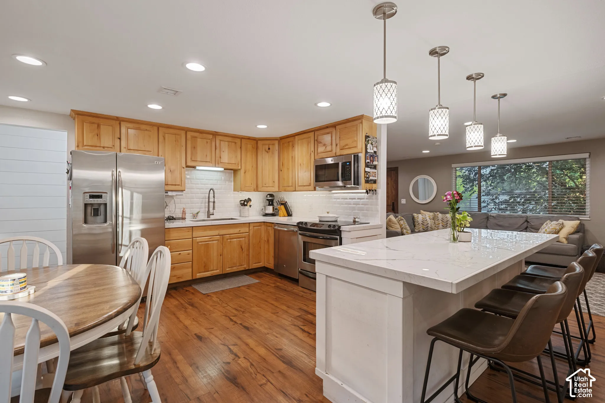 Wood-finish flooring throughout, stainless steel appliances, shaker-style wood cabinetry, subway tile backsplash, and a large island with a light-toned countertop
