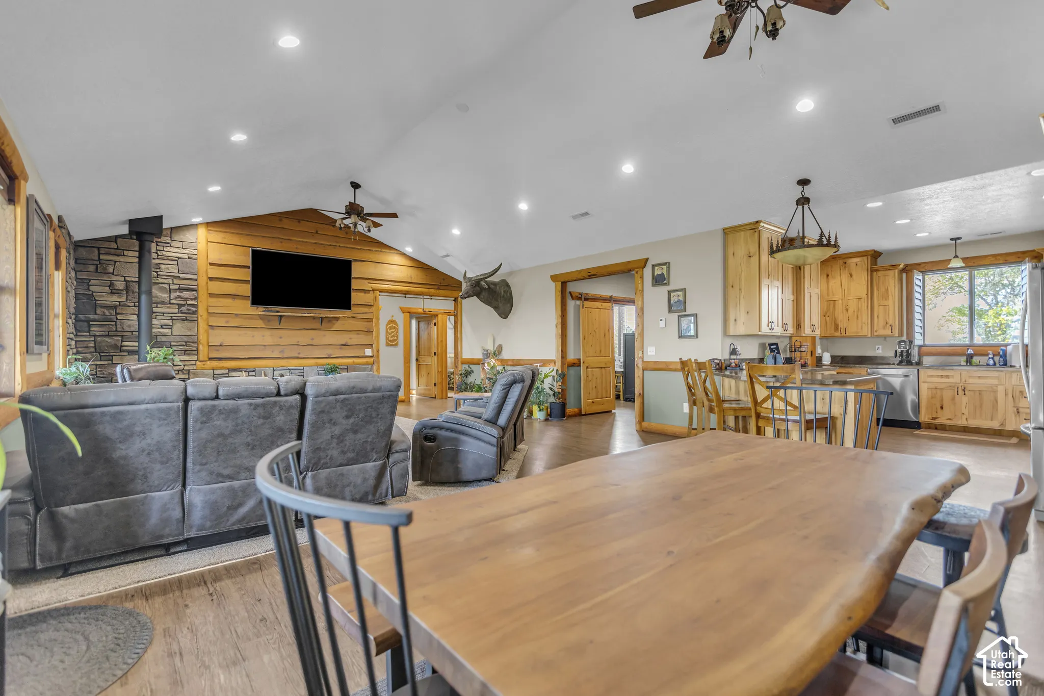 Vaulted ceiling with recessed lighting, stone-wrapped wood-burning stove, wood-finish flooring, natural wood cabinetry, and stainless steel appliances