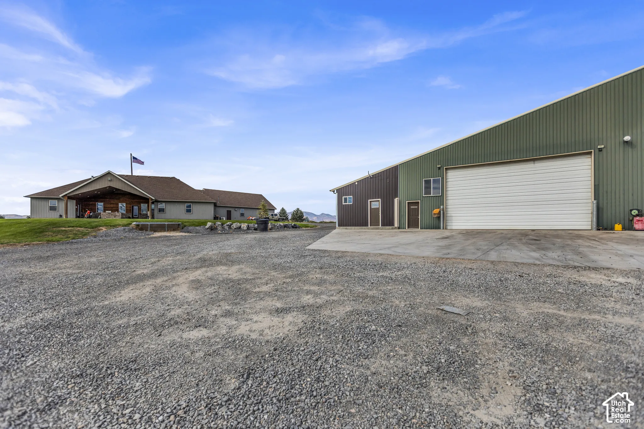 Gravel driveway with a multi-story house featuring a covered front porch, and a large outbuilding with a roll-up door and multiple entry points