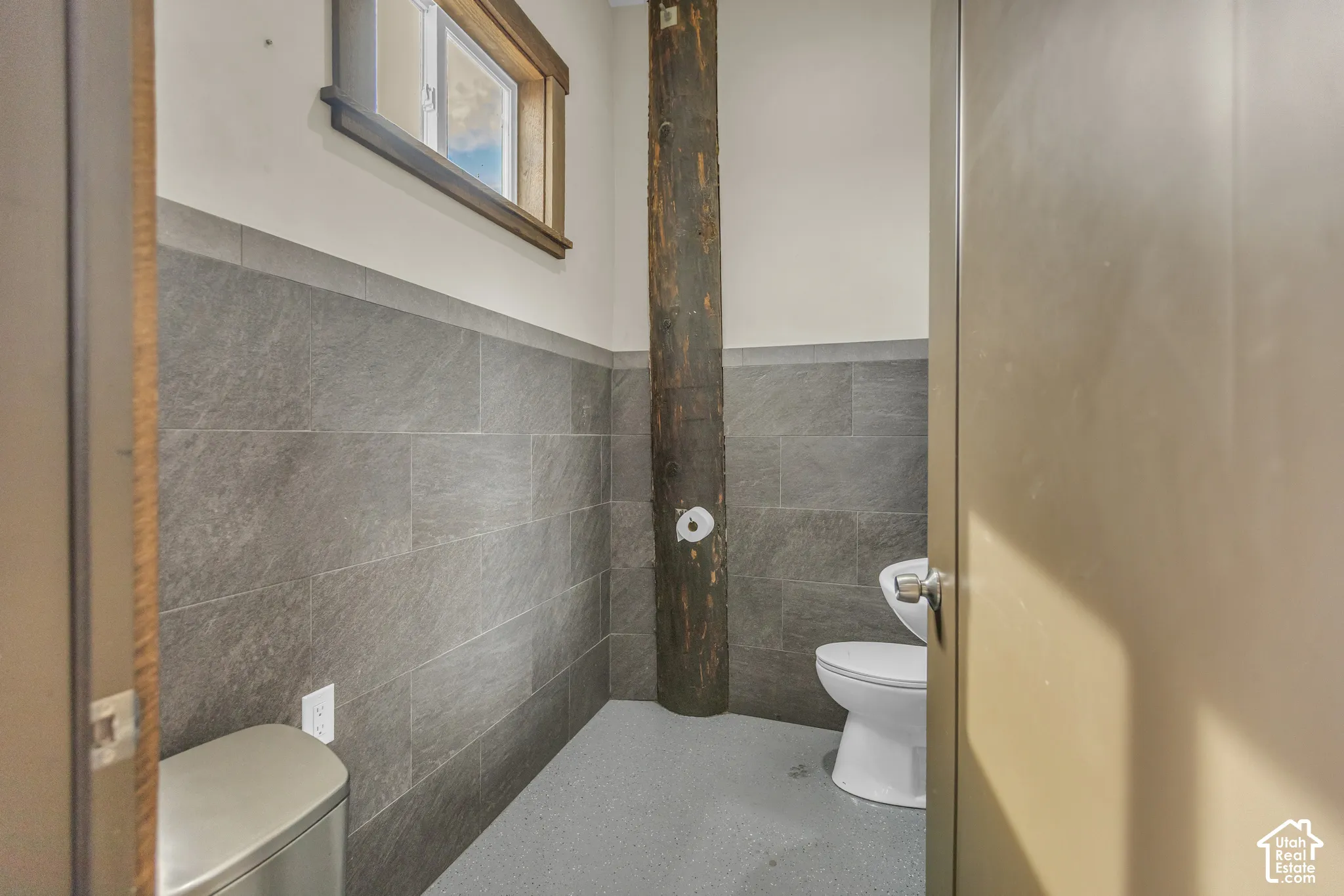 Half-wall tile wainscoting, a rustic exposed wood beam, a window with wood trim, and a white toilet with a wall-mounted paper holder