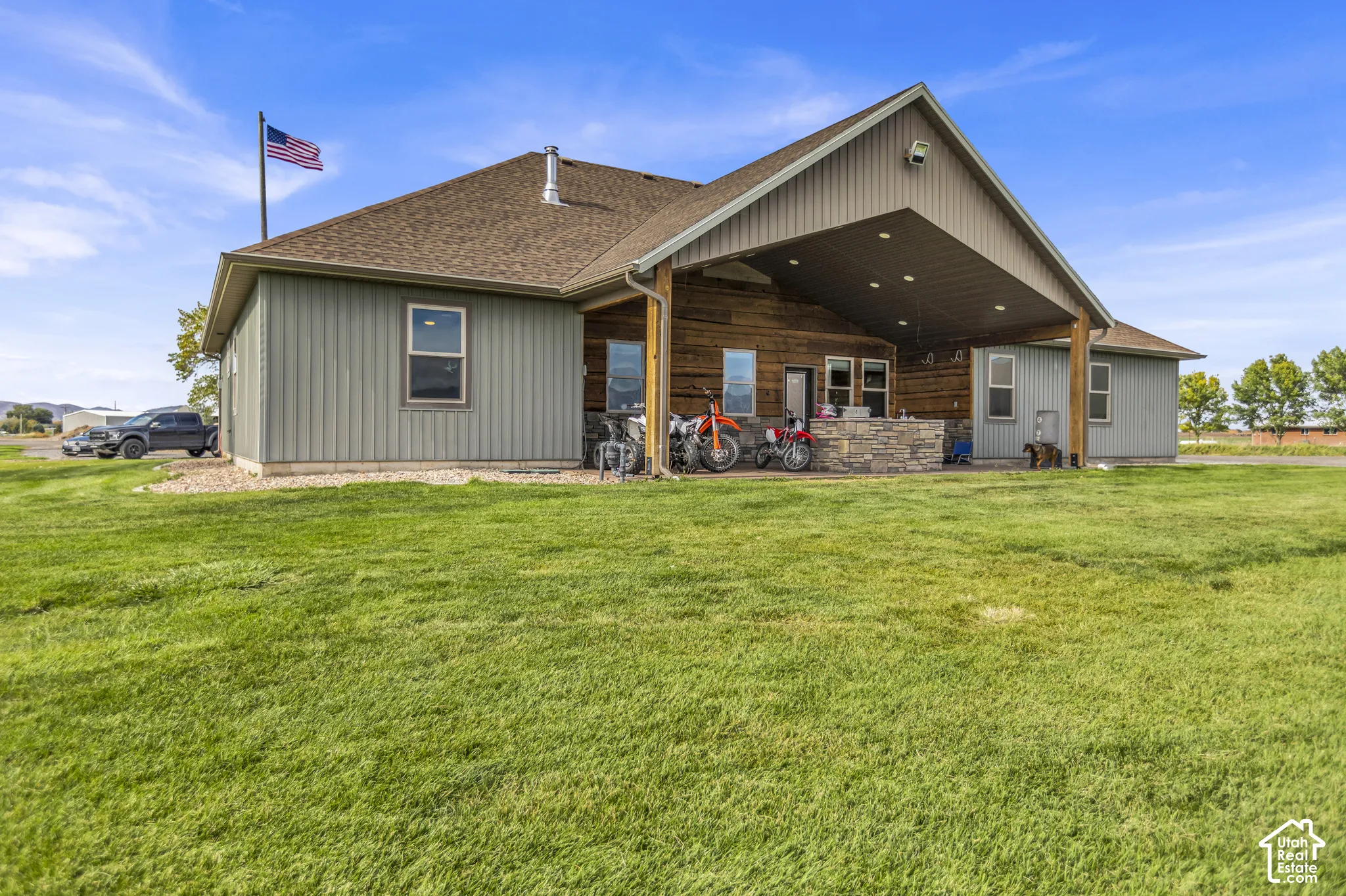 Expansive green lawn, covered outdoor patio, stone-clad outdoor kitchen, vertical siding, and a gabled roofline