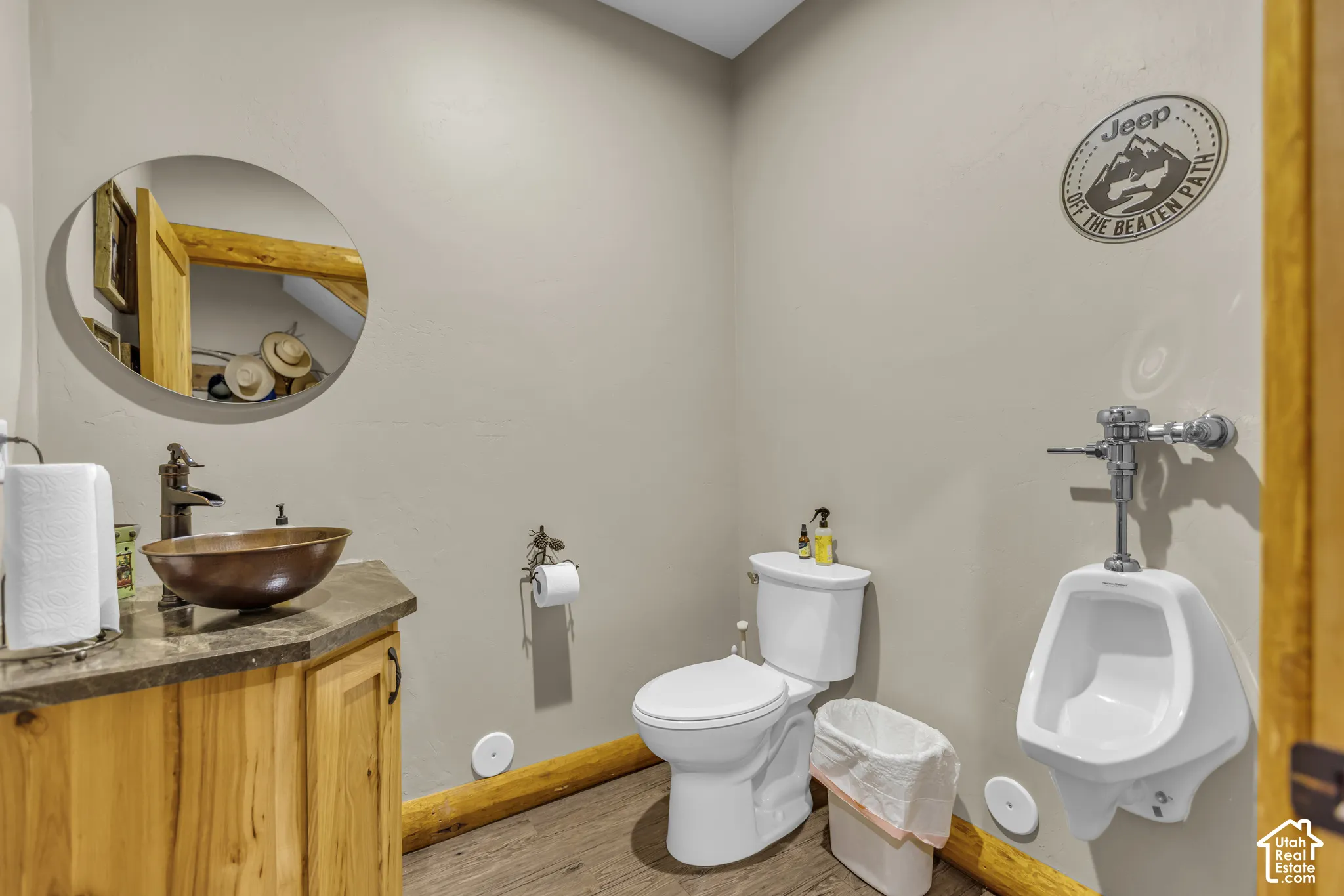 Wood-finish flooring with a toilet, urinal, and wood vanity with a vessel sink and a circular mirror