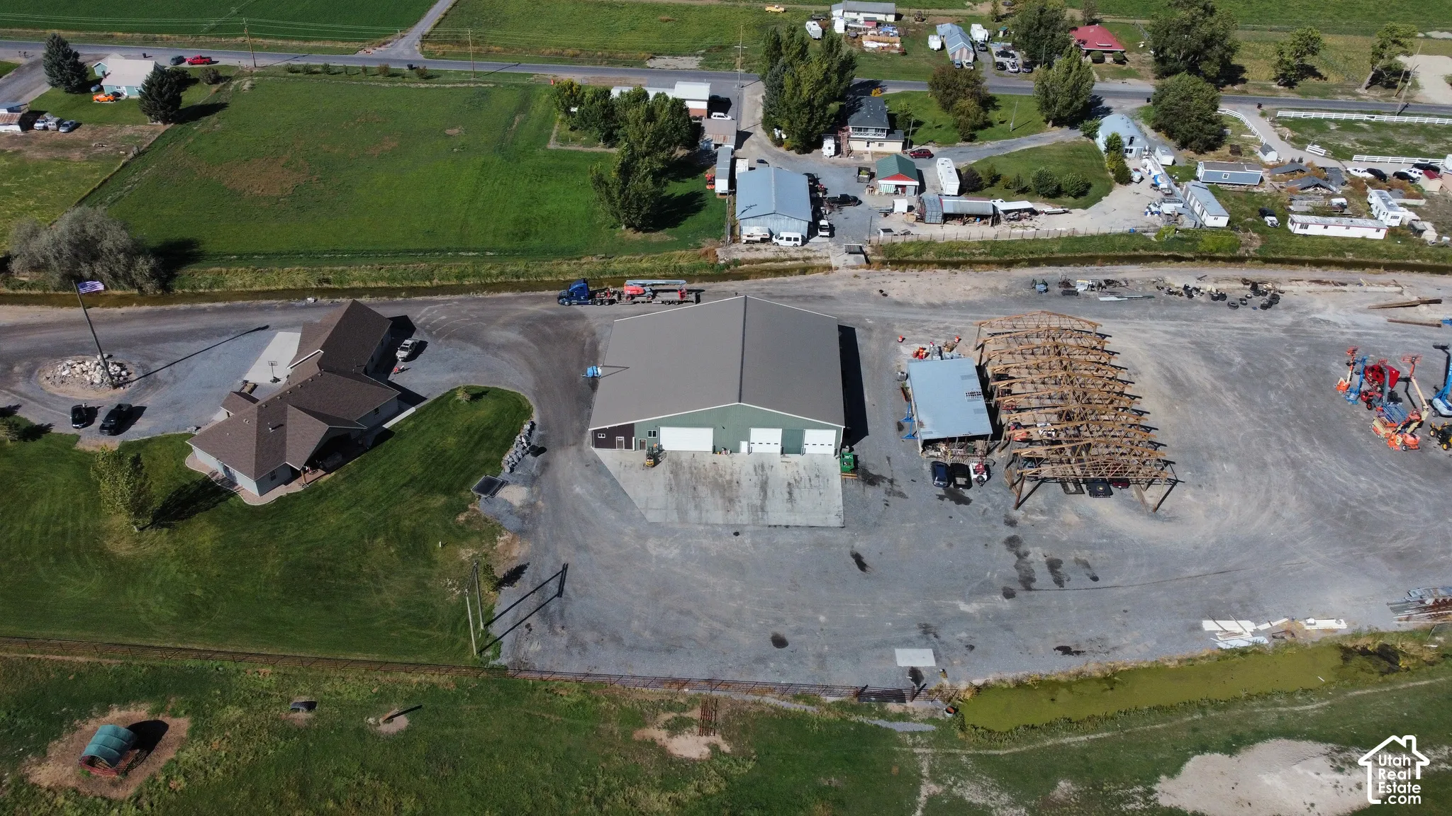 Expansive gravel lot with multiple structures, including a two-story building with a brown shingle roof, a large commercial-style building with a gray roof and green doors, and an open-sided shed with a light-colored roof