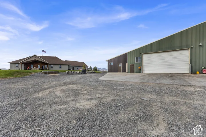 Gravel driveway with a multi-story house featuring a covered front porch, and a large outbuilding with a roll-up door and multiple entry points